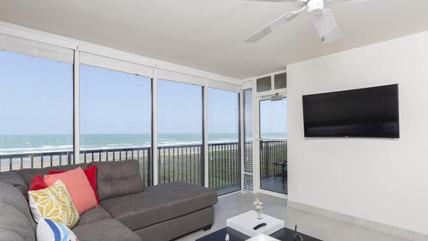 Ein Balkon mit Meerblick, ein graues Sofa, ein an der Wand befestigter Fernseher und ein Deckenventilator.