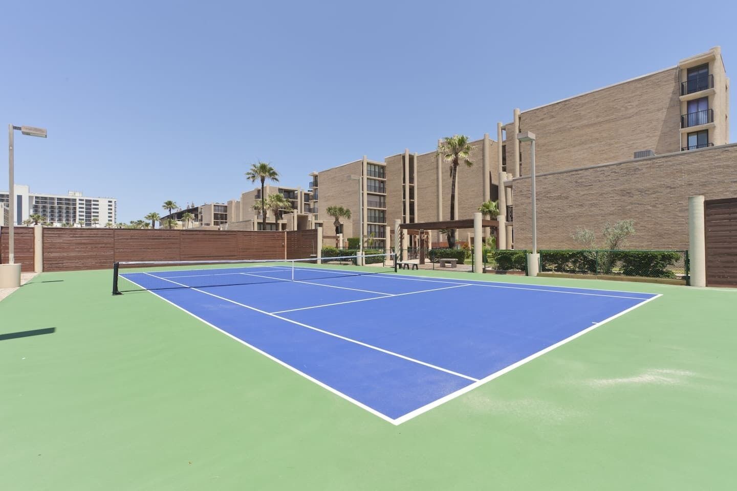 A tennis court with a net, surrounded by a wooden fence and a brick building with balconies.