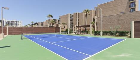 A tennis court with a net, surrounded by a wooden fence and a brick building with balconies.