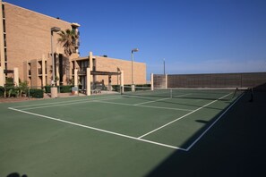 Una cancha de tenis con red, rodeada por un edificio con varios pisos y balcones.