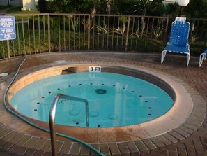 Un jacuzzi rond avec un escalier en métal, entouré d’une terrasse en briques, et une chaise bleue en arrière-plan.