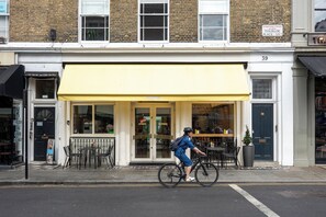 A person riding a bicycle past a building with a yellow awning and a black door. - Modern Split-Level flat, South Kensington, London - Pass the Keys (London)