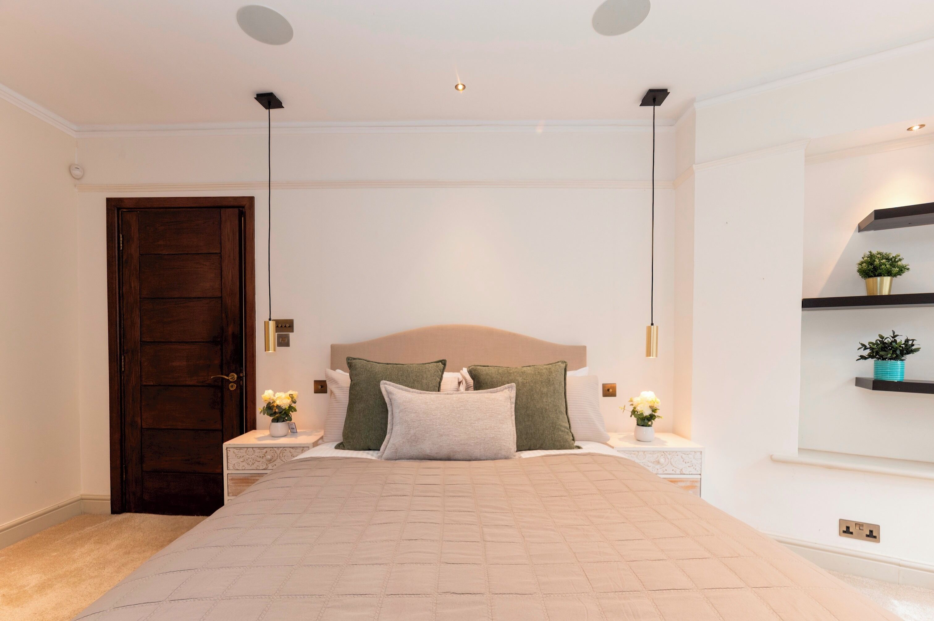 A bedroom with a large bed, bedside tables, a wooden door, and a wall-mounted shelf with a plant.