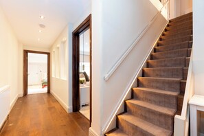 A hallway with wooden flooring, a staircase with carpeted steps, and a door leading to another room. - Luxury Family Home with Garden in West Kensington - 2 Mins to Tube - Pass the Keys (Londonderry)