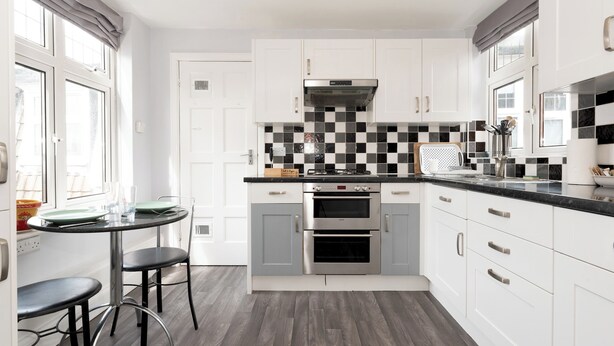 A modern kitchen with white cabinets, a black and white checkered backsplash, stainless steel appliances, and a round dining table with bar stools. - Riverside Serenity near Kingston and Hampton Court - Pass the Keys (Surbiton)