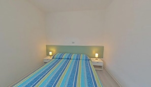 Ein kleines Zimmer mit einem Einzelbett, einer gestreiften Bettdecke, zwei Nachttischen mit Lampen und einer schlichten Wand.