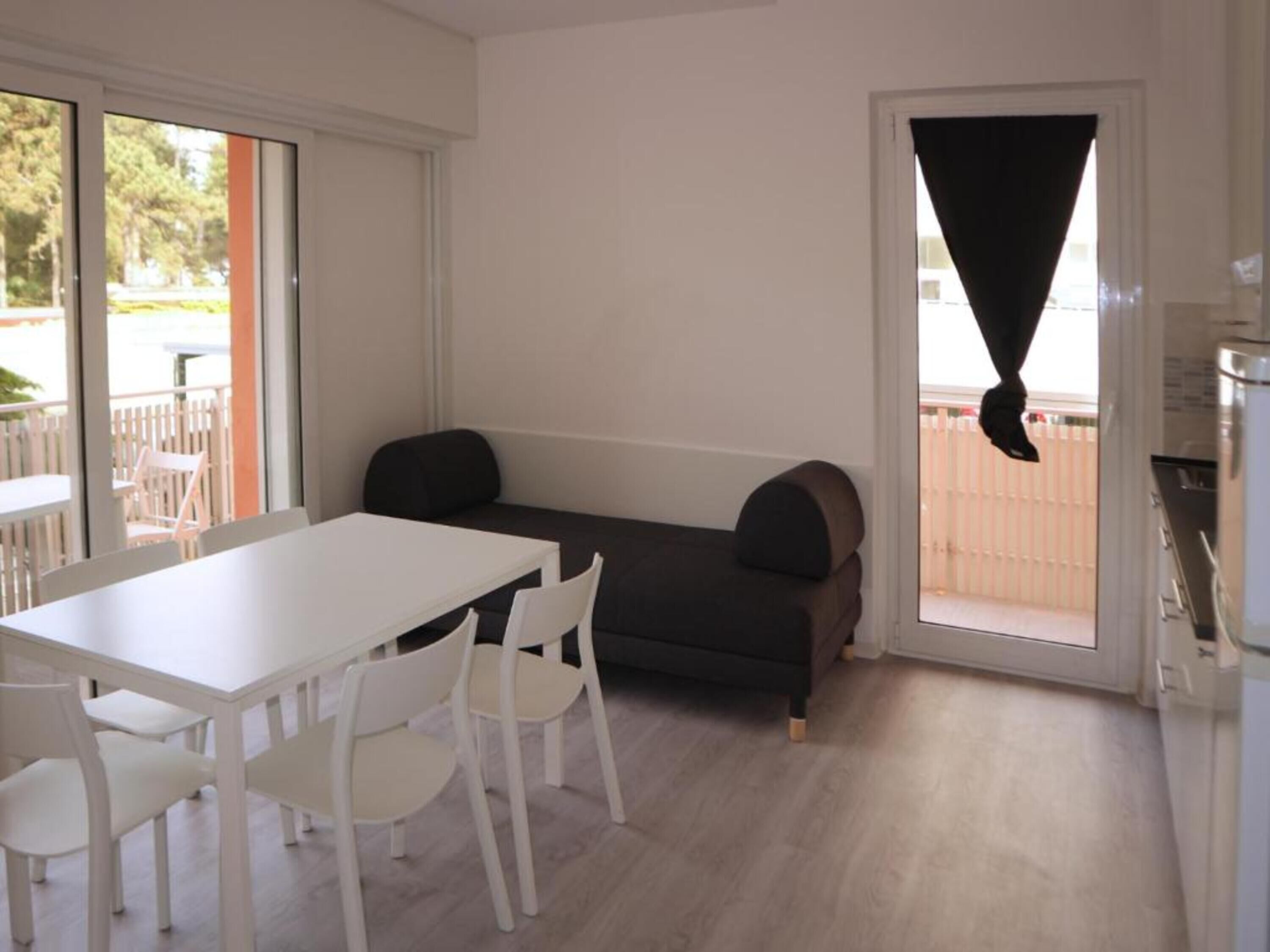 A room with a white table, four white chairs, a dark sofa, and a sliding glass door with a black curtain.
