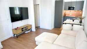 A modern living room with a flat-screen TV, a sofa, a wooden entertainment unit, and a kitchen area with a dining table. - Renovated 2-room apartment 30 m from the beach - Le Mourillon (Toulon)