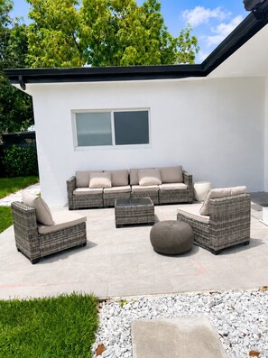 An outdoor patio with wicker furniture, a glass coffee table, and a large window. - Luxury Miami Retreat w/ Beautiful Backyard | 3BR (Miami)