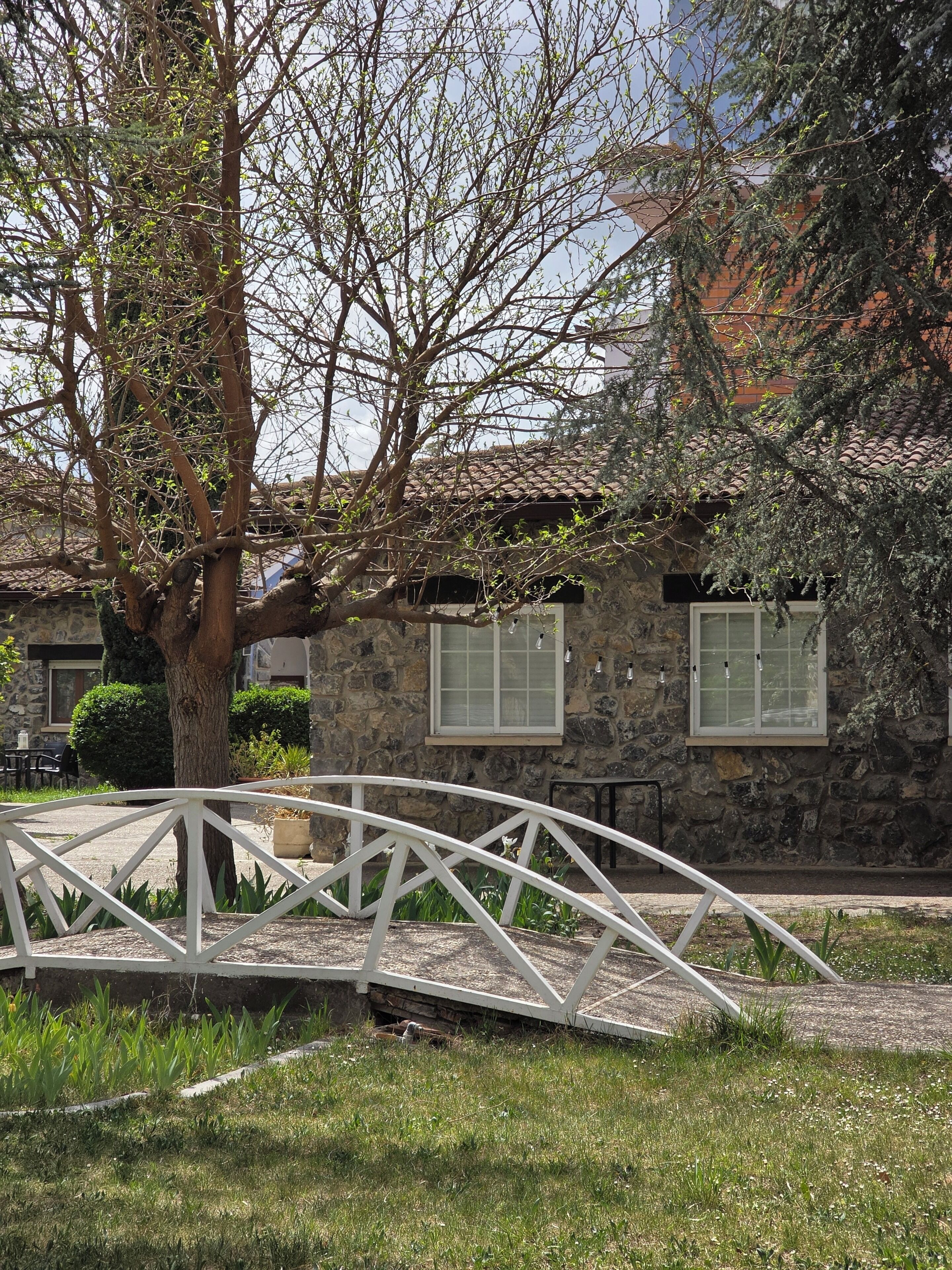 Un edificio de piedra con techo de tejas, un puente de metal blanco y un árbol en primer plano.