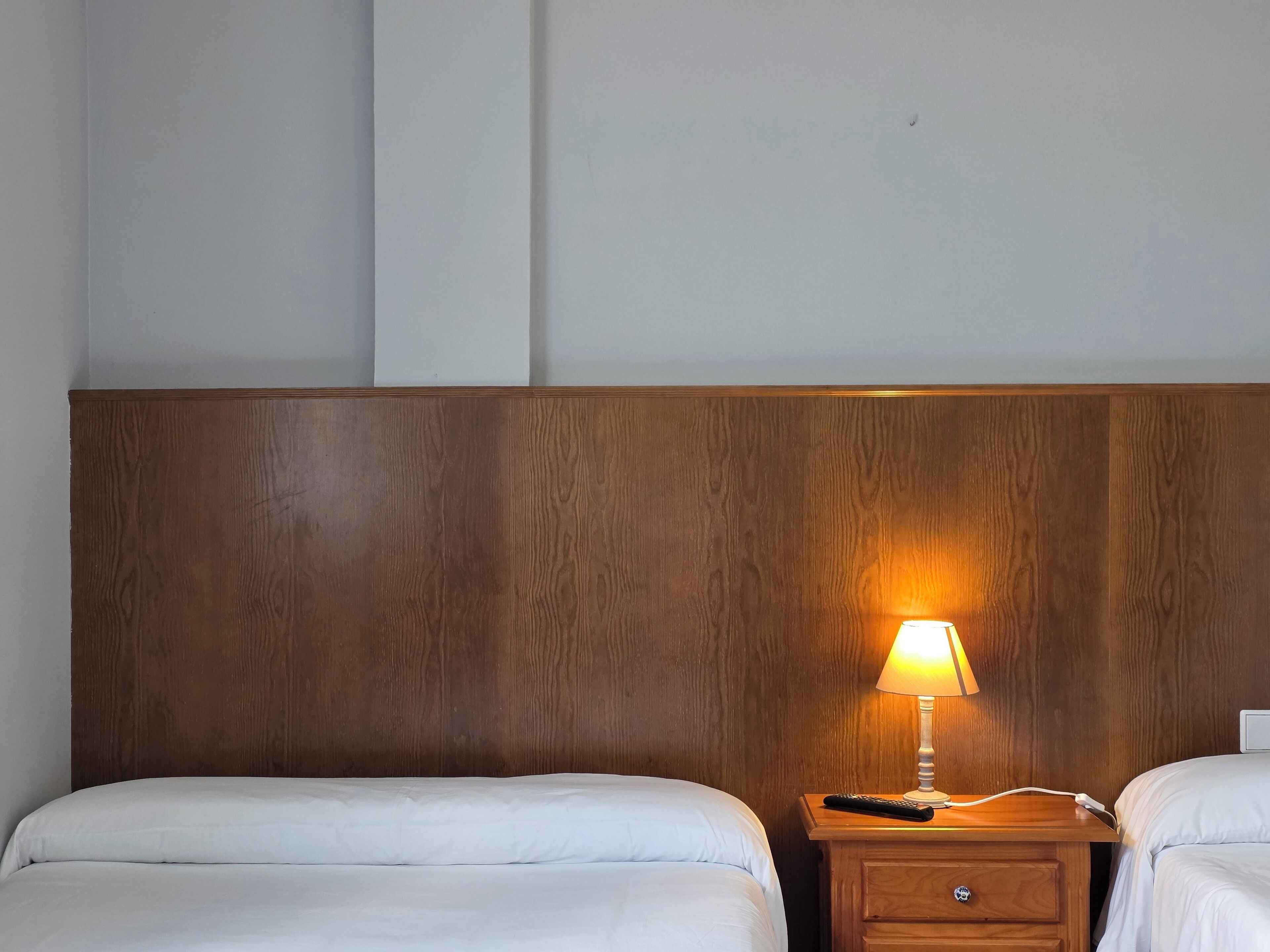 Habitación de hotel con dos camas, cabecera de madera, mesita de noche con lámpara y un cajón.