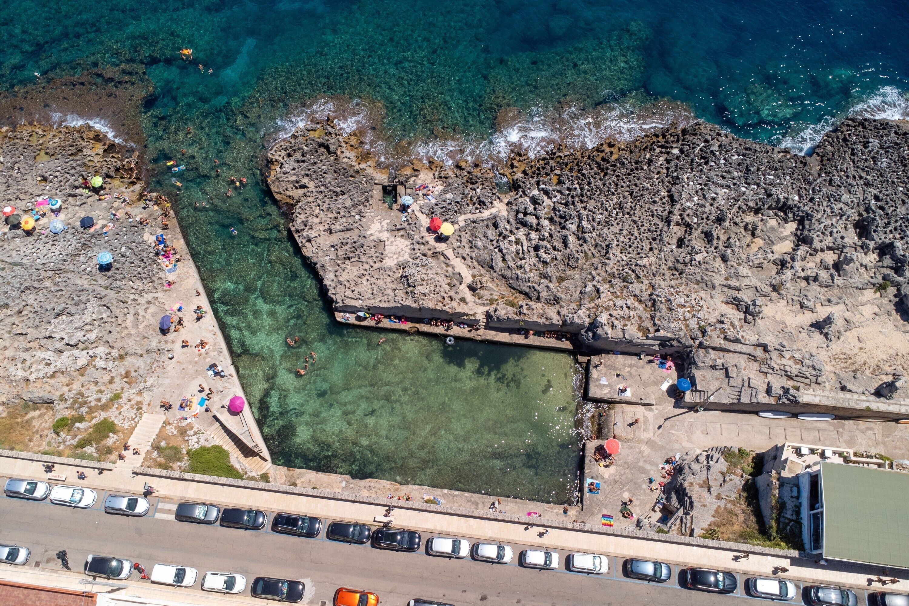 An aerial view of a coastal area with rocky terrain, clear water, and a paved road with parked cars.