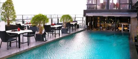 An outdoor pool area with tables and chairs, a bar, and a view of the city.