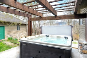 Un jacuzzi bajo una pérgola de madera con una pared de piedra y una casa al fondo.