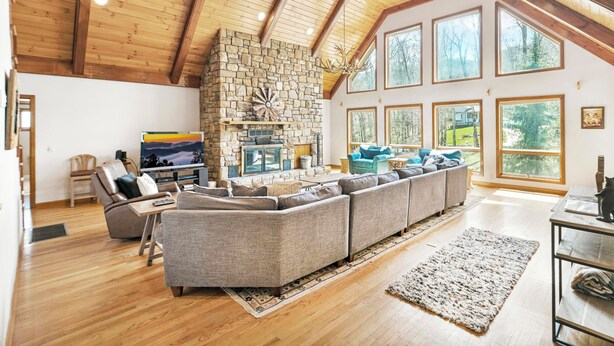 A spacious living room with a stone fireplace, large windows, and a high wooden ceiling.