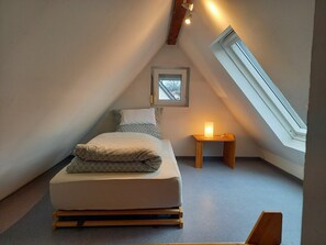 Een kleine, knusse kamer onder het dak, met schuine wanden, een enkel bed, een nachtkastje met een lamp en een raam dat natuurlijk licht biedt.