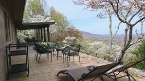 Um deck de madeira com móveis de exterior, incluindo cadeiras e uma mesa, com vista para uma paisagem deslumbrante com árvores e montanhas.