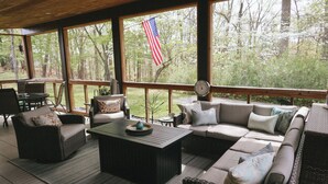 Uma área de estar coberta ao ar livre com móveis de vime, uma mesa de centro e vista para árvores e uma bandeira americana.