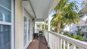 Een houten balkon met witte leuningen, een tafel met stoelen, en uitzicht op omliggende huizen en palmbomen.