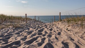 Ein Sandweg am Strand, der zum Meer führt, mit Holzpfählen und vereinzelter Vegetation.