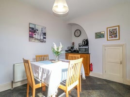 Un comedor con una mesa, sillas, un jarrón de flores, un reloj y una zona de cocina con microondas.