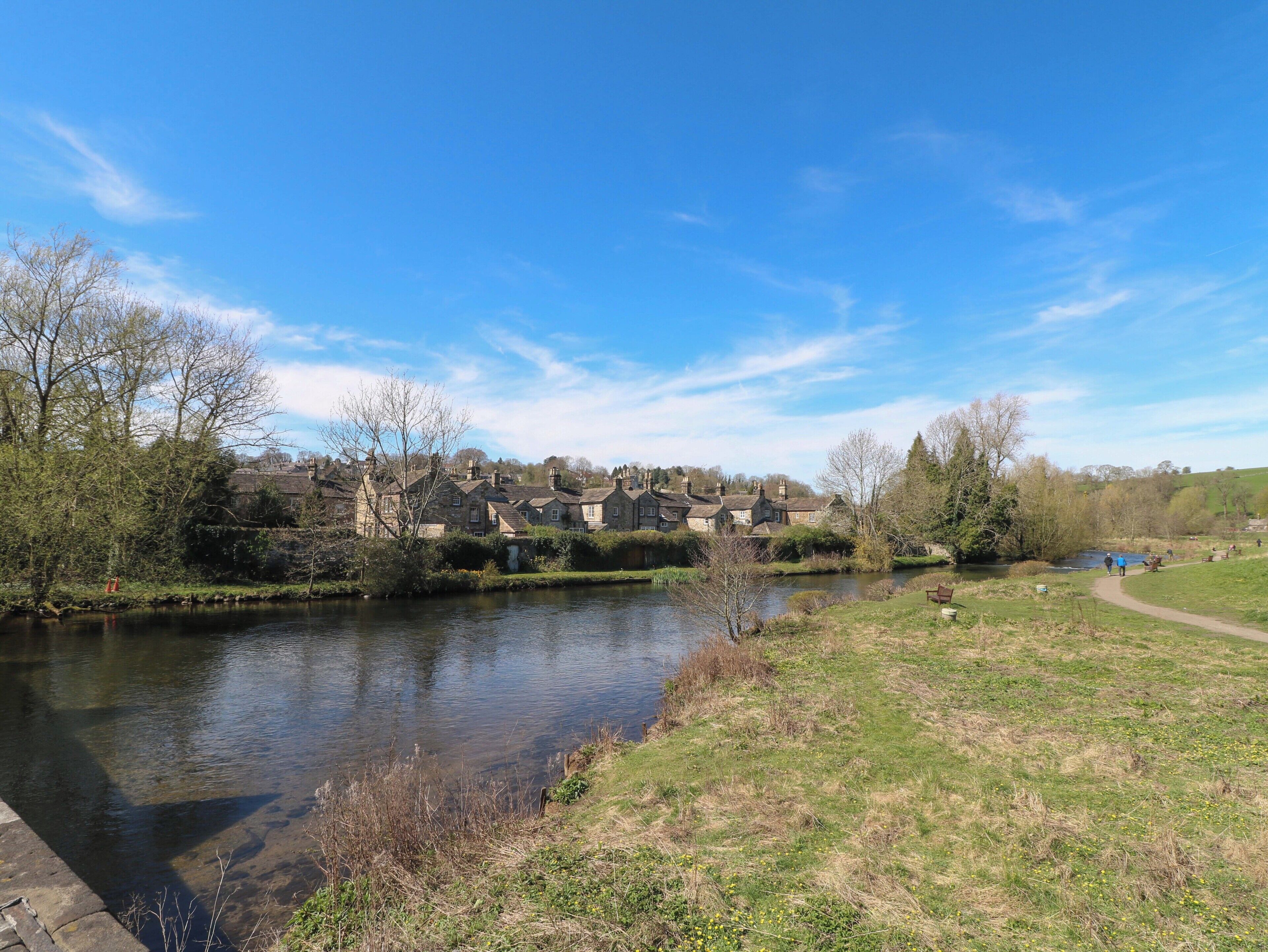 Blick auf einen Fluss mit Steinhäusern, einem Weg und viel Grün.