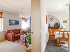 A living room with a sofa, a dining table with chairs, a kitchen area with appliances, and a clock on the wall.