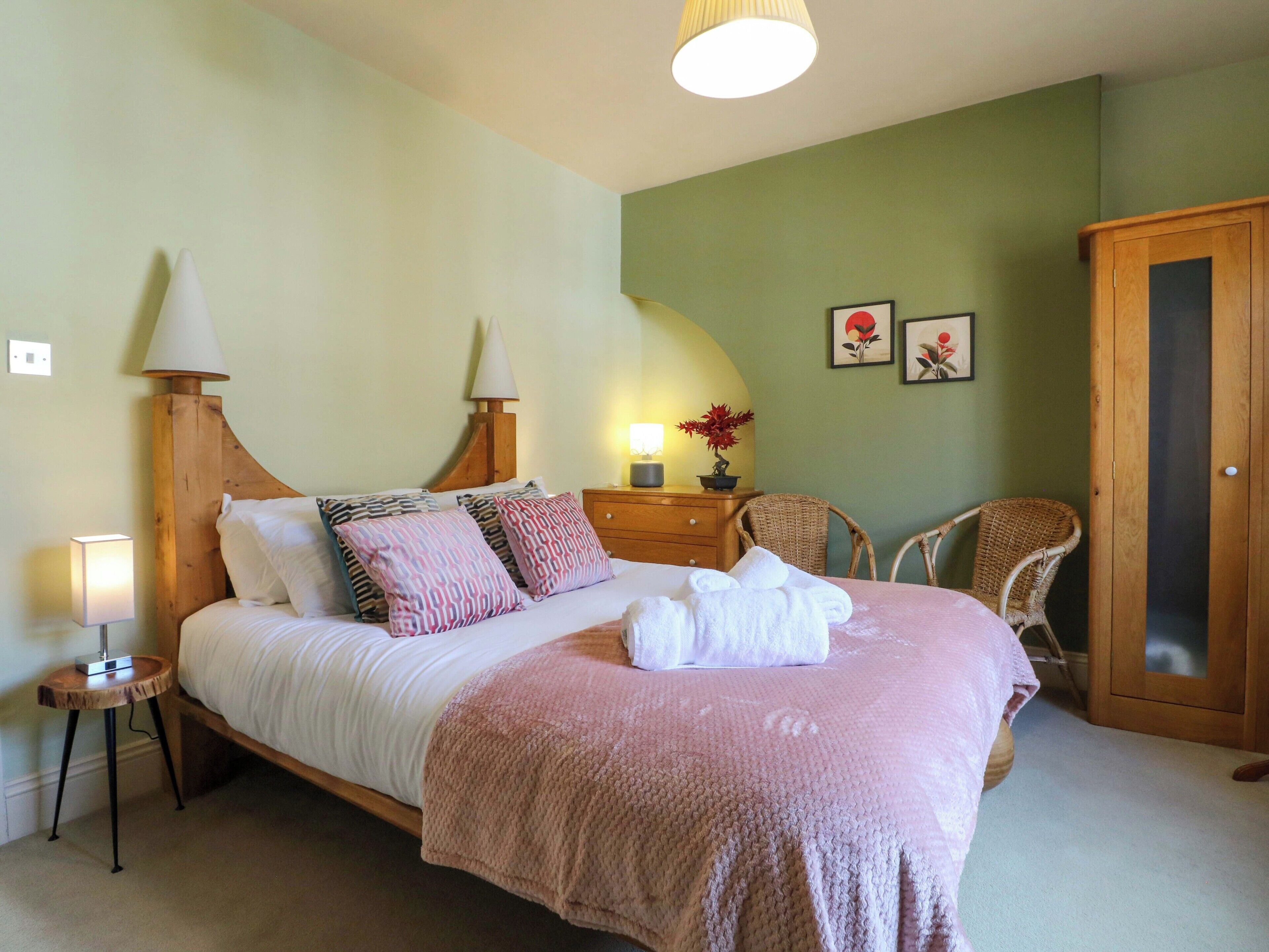 Una cama bien hecha con almohadas, una lámpara de noche, una mesita con lámpara, una silla y un armario en una habitación con paredes verdes.