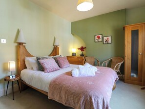 A neatly made bed with pillows, a bedside lamp, a small table with a lamp, a chair, and a wardrobe in a room with green walls. - 2A Water Street (Bakewell)