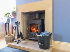 Una habitación con chimenea, una cesta, un cubo metálico con la etiqueta 'ASH' y una mesa cubierta con un mantel azul.