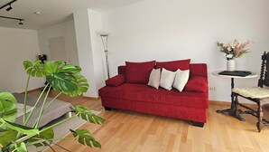 Un canapé rouge avec des coussins blancs, un grand lampadaire, une petite table sur laquelle est posé un vase de fleurs et une chaise en bois avec un coussin.