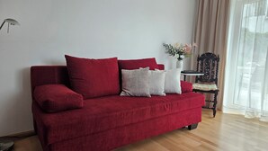 Un canapé rouge avec des coussins, un vase avec des fleurs, une chaise et une lampe dans une pièce dont le sol est en bois et qui possède une fenêtre avec des rideaux.