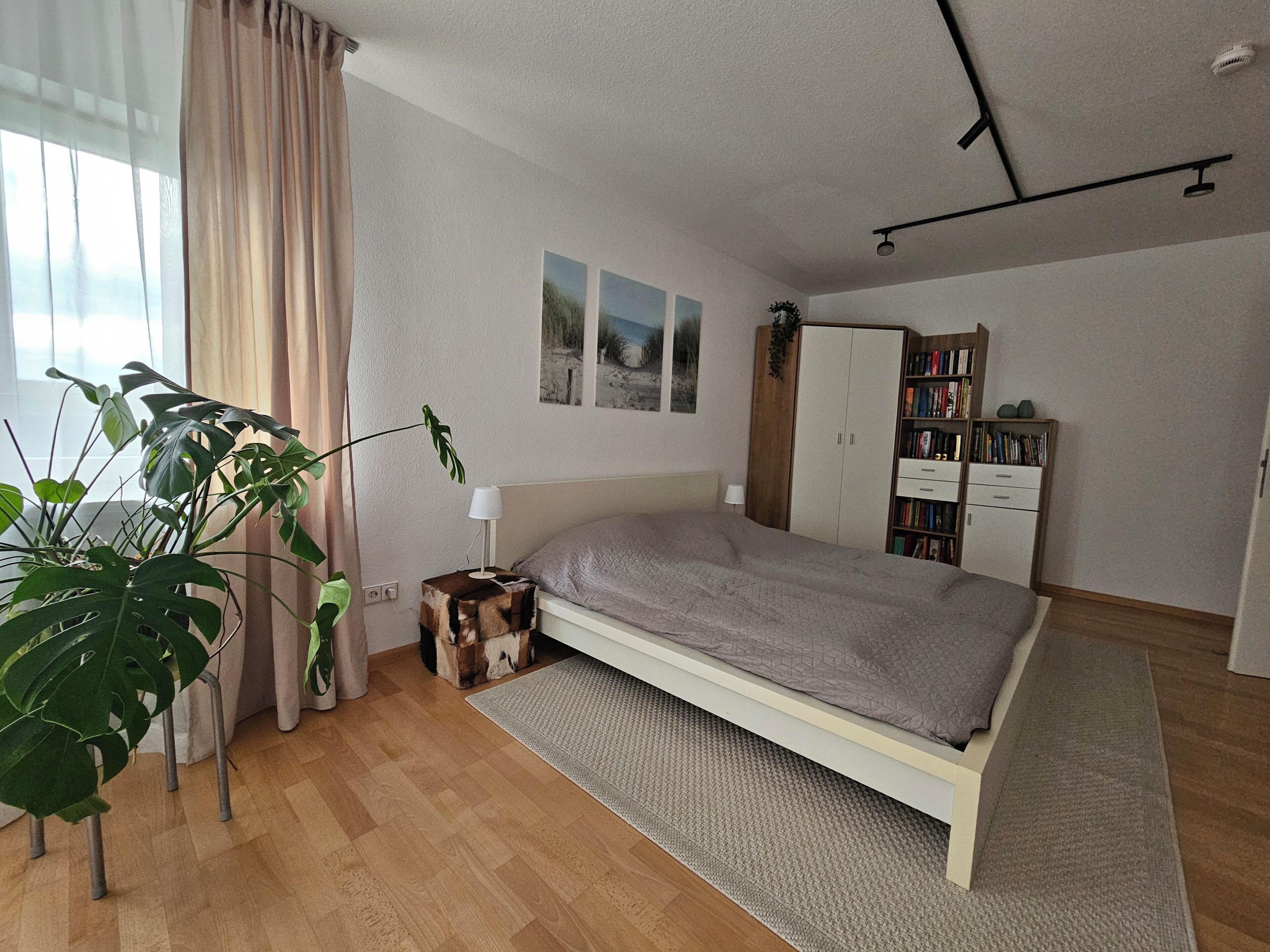 Ein Schlafzimmer mit Bett, Kleiderschrank, Schreibtisch, einer Zimmerpflanze und einem Fenster mit Vorhängen.