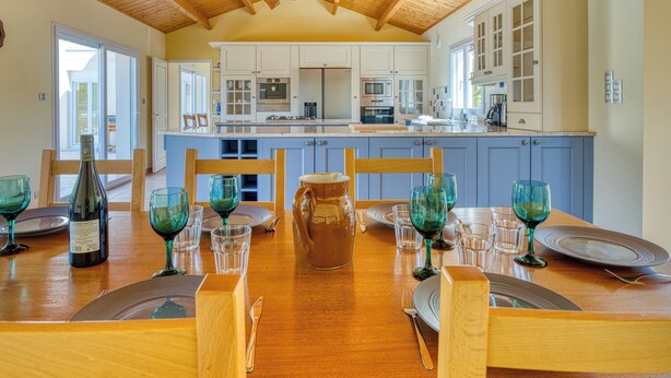 A dining area with a wooden table set for six, a large wooden ceiling, and a kitchen in the background.
