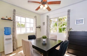 Une pièce dotée d’un ventilateur de plafond, d’un distributeur d’eau, d’une table avec des chaises et de grandes fenêtres avec des rideaux.