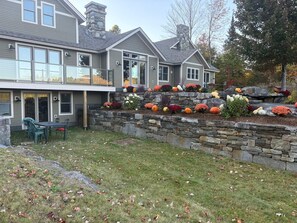 Une maison à deux étages, dotée d’un mur de soutènement en pierre, d’un patio couvert avec des chaises et d’une variété de feuillages aux couleurs vives, typiques de l’automne.