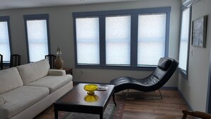 A living room with a sofa, a coffee table, a black leather chair, and large windows with blinds.