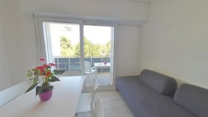 A room with a white table, a grey sofa, and a sliding glass door leading to an outdoor area with a table and chairs. - Side Sea-view in Lignano - 30 m From the Beach (Lignano Sabbiadoro)