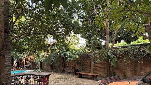 A hammock, a wooden table, and a bench in a garden with trees and a brick wall. - 403 Serene Teak Boutique Apartments (Chiang Mai)
