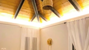 A room with a sloped ceiling, a bed, a dresser, a wicker chair, a window with curtains, and a wall-mounted light fixture.