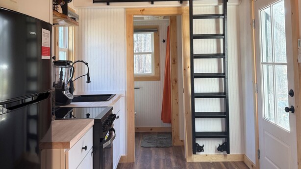 A kitchen with a ceiling fan, a black refrigerator, a toaster, and a wooden floor. - Tiny House in the Woods (Oakland)