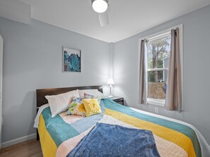 Ein Schlafzimmer mit einem Bett, einem Nachttisch mit Lampe, einem Fenster mit Vorhängen und einem an der Wand hängenden Kunstwerk.