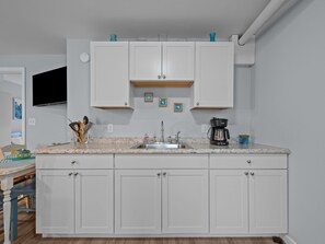 A kitchen with white cabinets, a granite countertop, a coffee maker, and a TV mounted above the cabinets. - Emerald Escape Down - 1BR - Short walk to beach! (Emerald Isle)