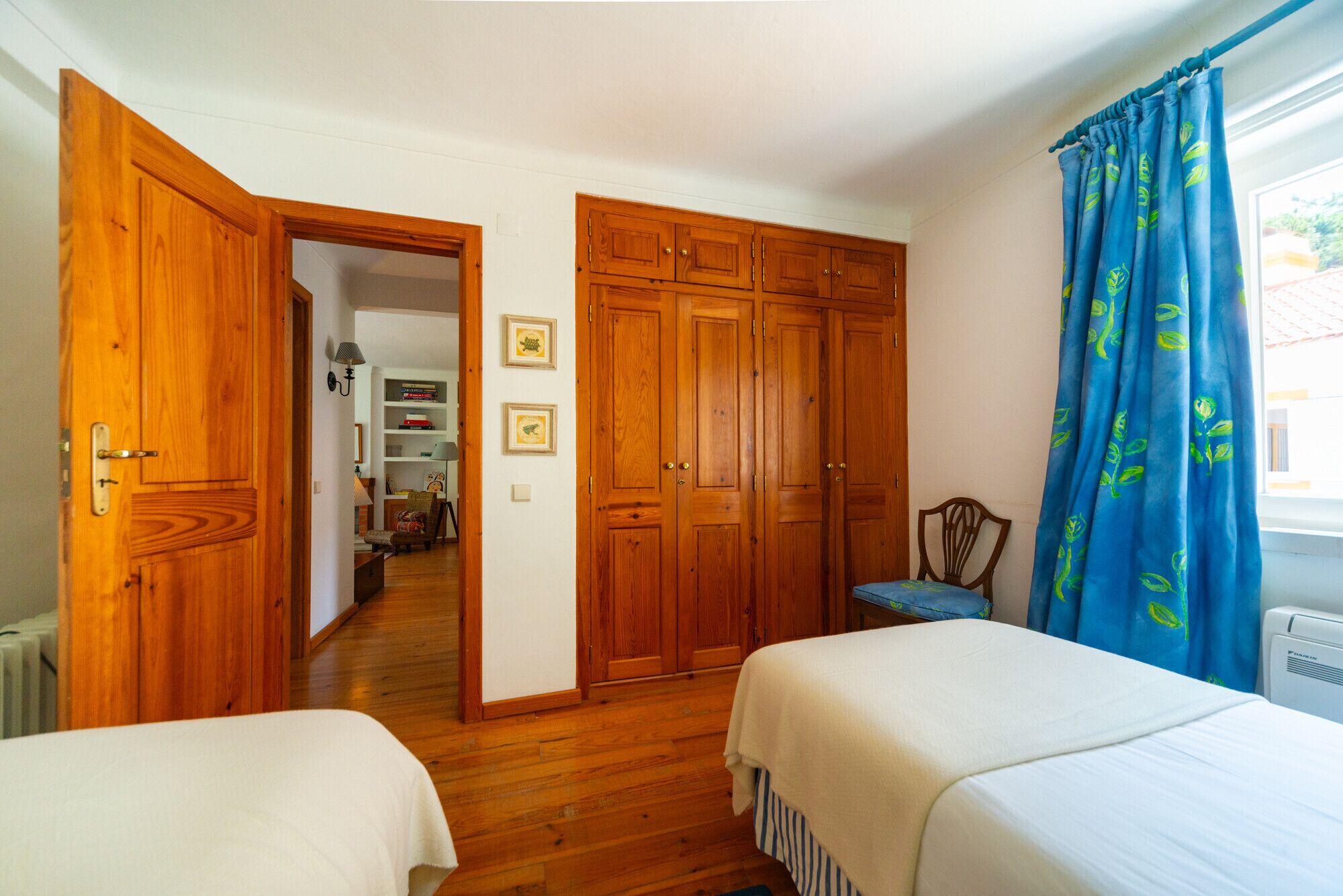 A bedroom with two beds, wooden wardrobe, and a window with blue curtains.