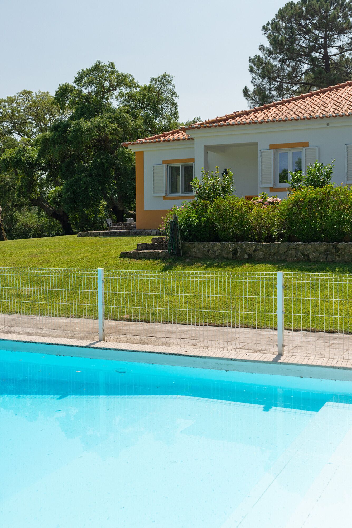 Une piscine entourée d’une clôture blanche, une maison avec un toit en tuiles et une pelouse avec des arbres.