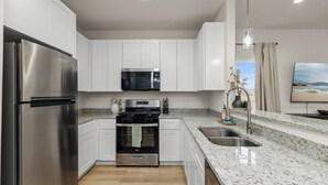 Cocina moderna con armarios blancos, electrodomésticos de acero inoxidable, encimeras de granito y un amplio ventanal con vista a la playa.