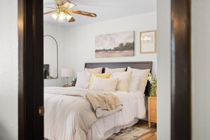 Quarto com cama, mesa de cabeceira, abajur, ventoinha no teto e obras de arte na parede.