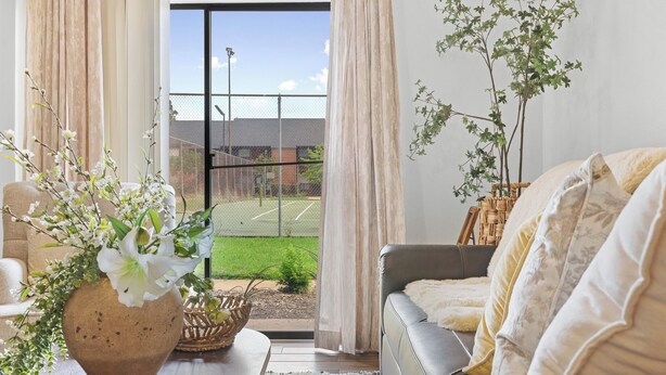 Ett vardagsrum med en soffa, ett soffbord med en vas med blommor och utsikt över en tennisbana genom en glasdörr.