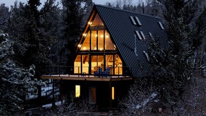 Una cabaña con techo inclinado, ventanas amplias y una terraza con sillas.
