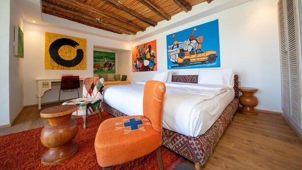 A bedroom with a bed, a chair, a desk, and artwork on the walls. - The House of Designers by BCKART GALLERY (Lalla Takarkoust)